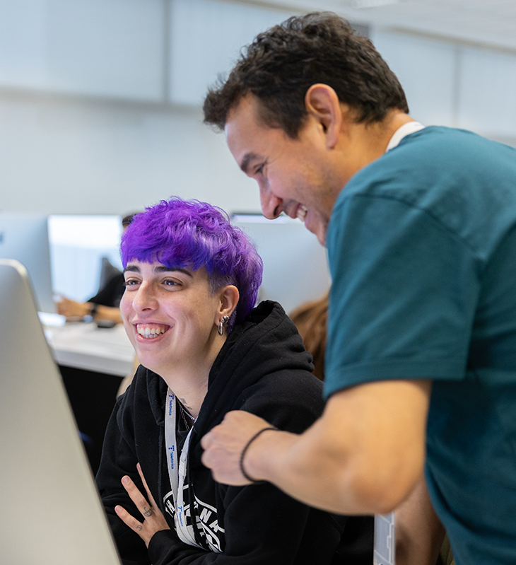 Person with purple hair smiles at a computer screen while a colleague in teal leans in to help.