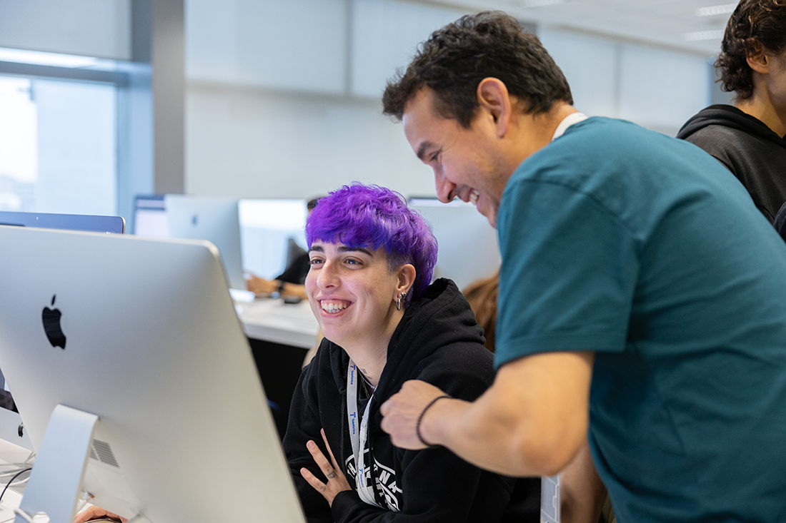 Dos personas colaboran frente a un ordenador en un espacio de trabajo de 42 Madrid Fundación Telefónica. Una está sentada utilizando el equipo mientras la otra permanece de pie junto a ella. Al fondo se observan otros ordenadores y personas trabajando.