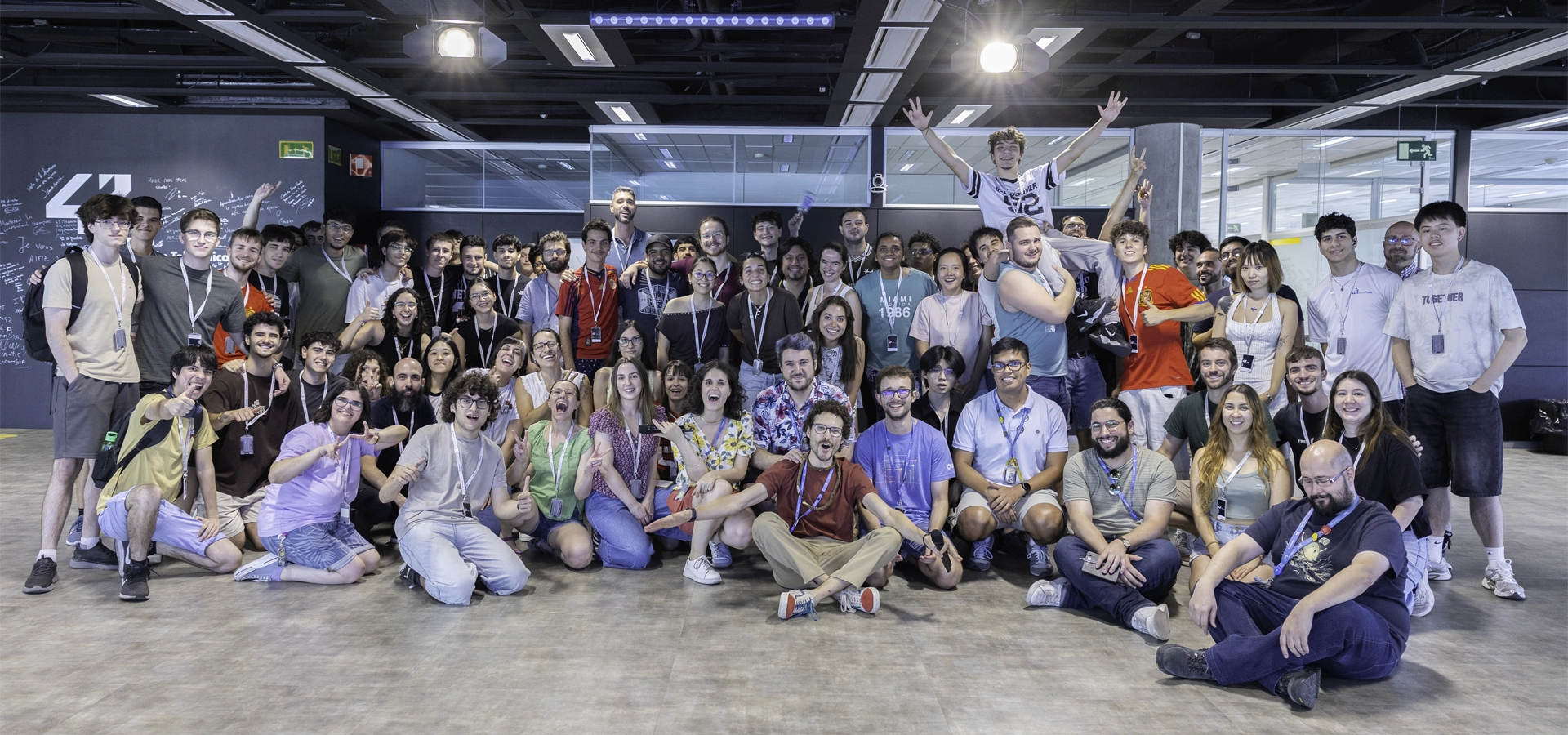 Large diverse group of people posing together in a bright, open-office environment, many wearing lanyards and smiling.",