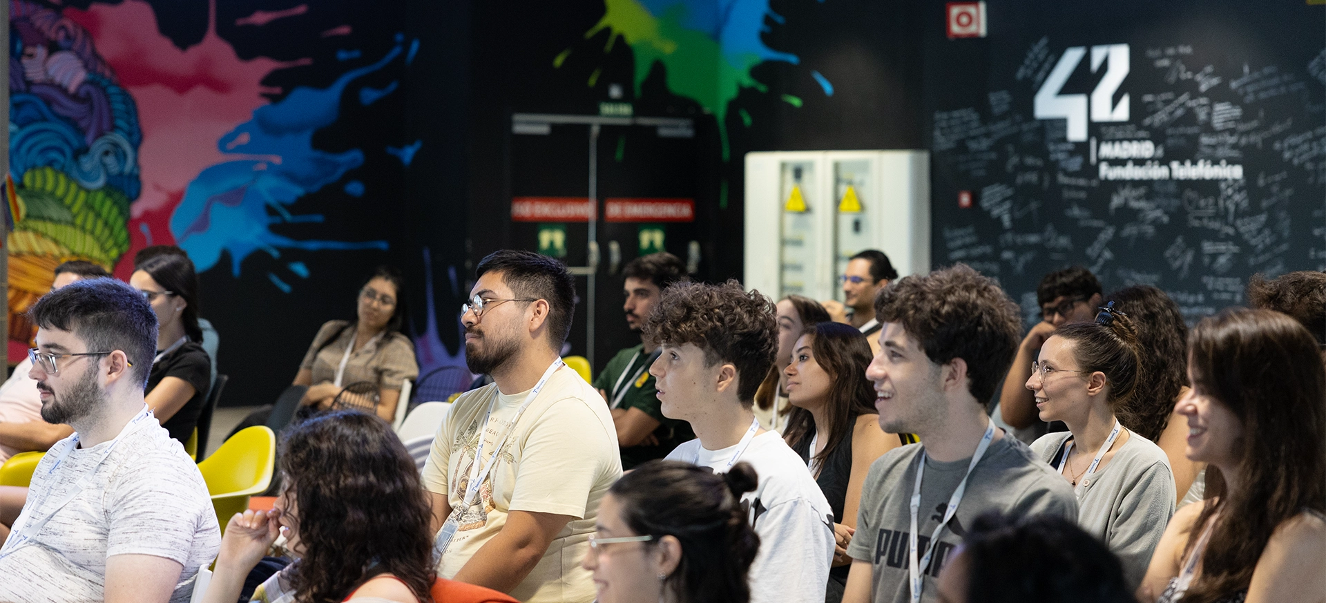 Diverse group of people seated and listening in a conference room with a colorful mural on the left and a signee wall on the right backdrop.