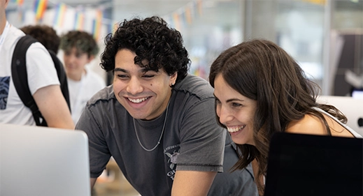 5 Two young people lean toward a laptop, smiling as they work together at an event or classroom setting.