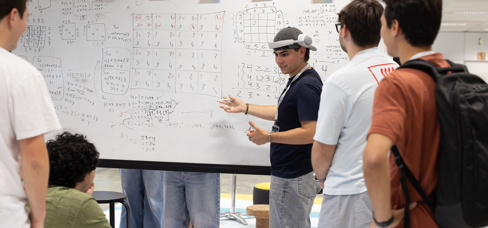 Group of students discusses math problems at a large whiteboard covered with grids and equations; one person gestures while explaining to peers