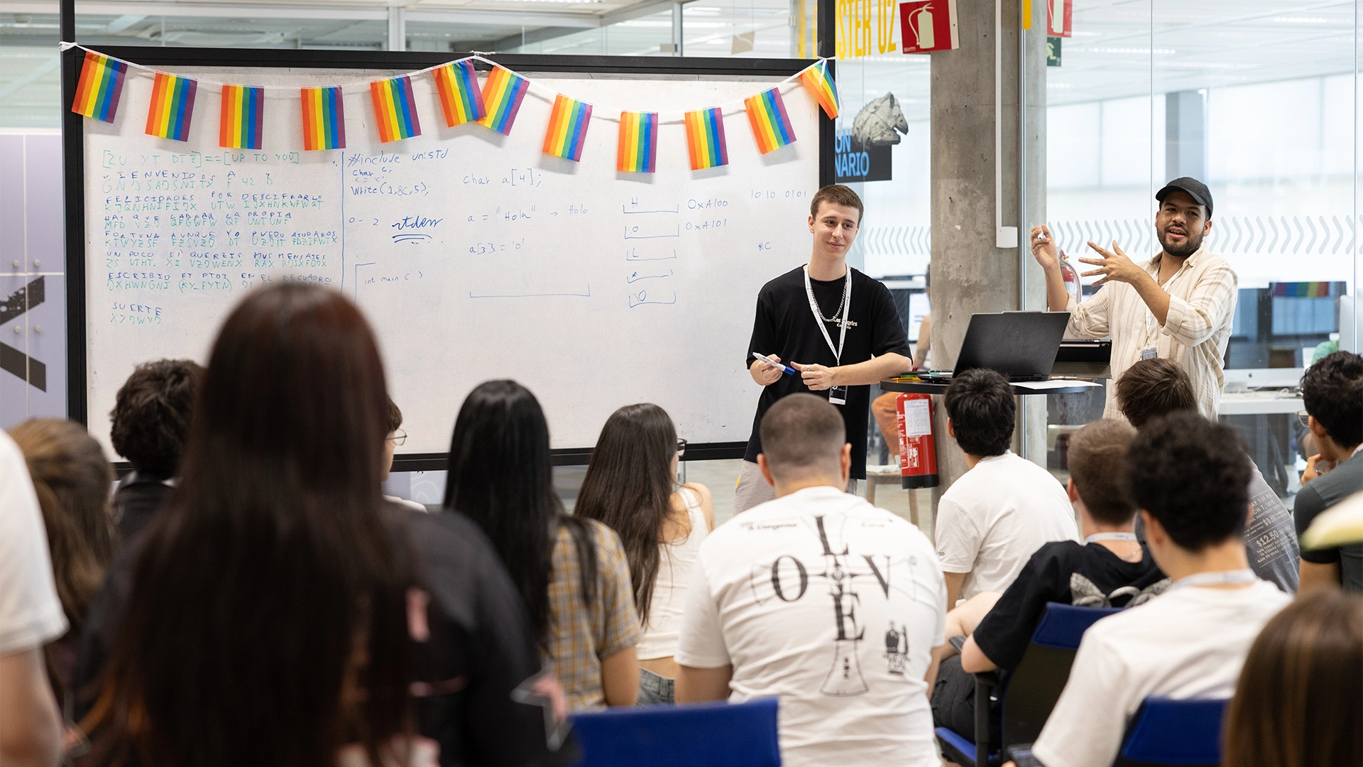 Two instructors present a workshop at the front of a classroom, writing on a whiteboard with colorful flags hanging overhead.