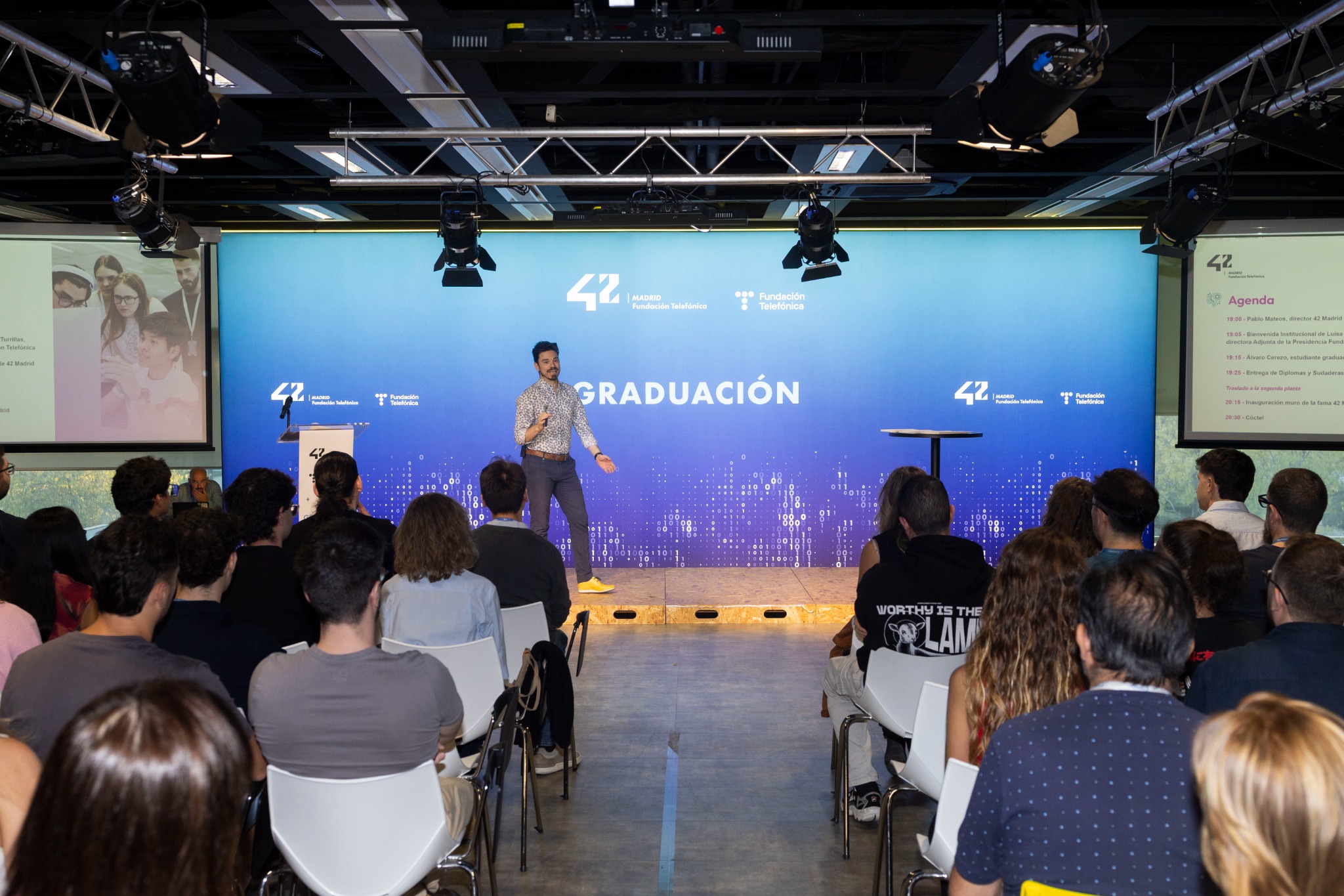 Speaker on stage gesturing to the audience during a graduation ceremony, with a blue backdrop showing 'GRADUACIÓN' and logos and people seated in front.
