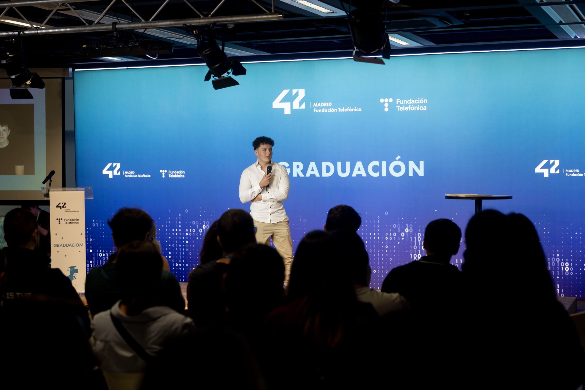 Male speaker on stage holding a microphone at a graduation ceremony, blue backdrop with 'GRADUACIÓN' and logos, audience in foreground.