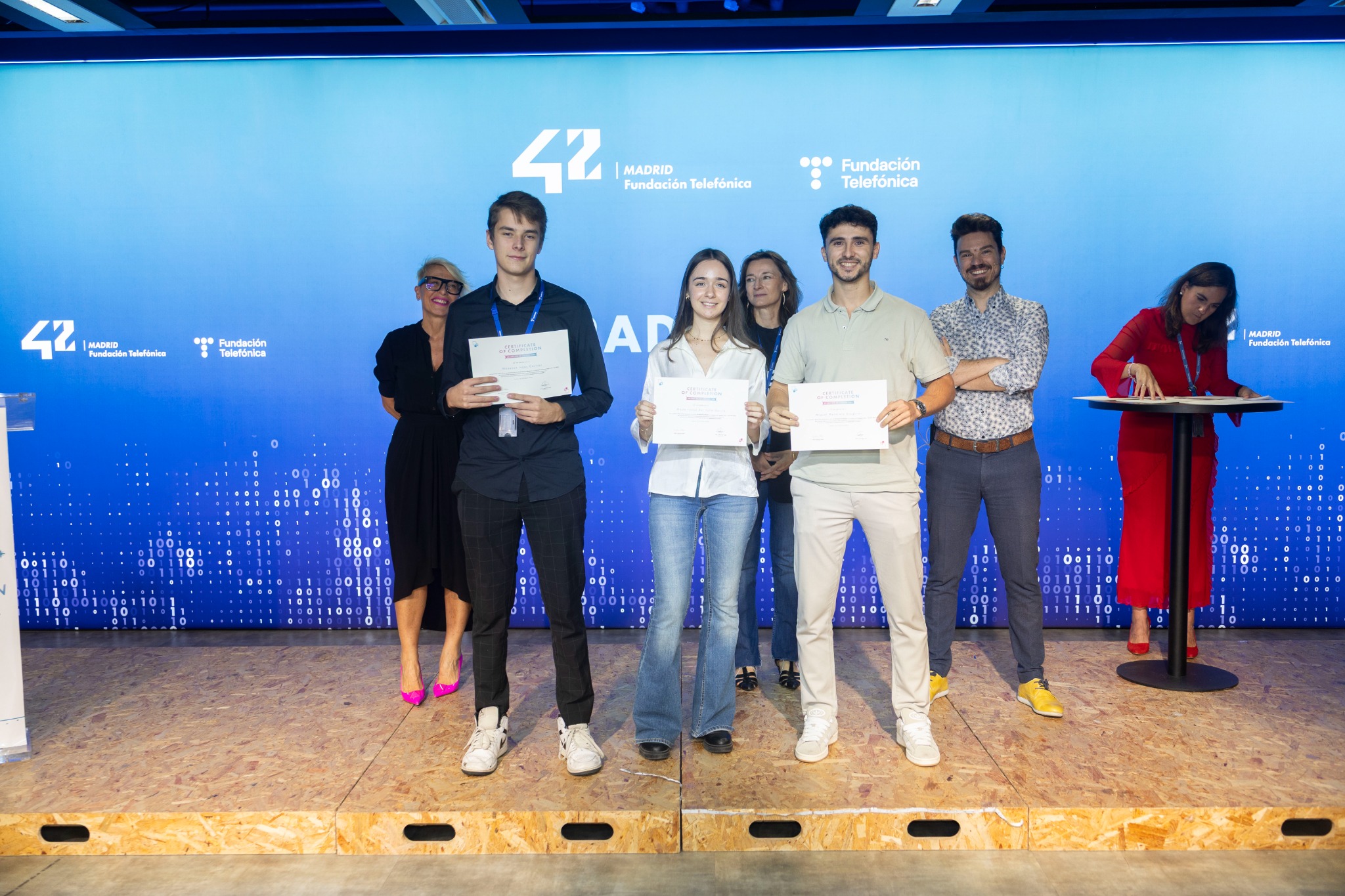 Group of six people on a stage holding certificates, posing for a photo in front of a blue backdrop with logos behind them.",