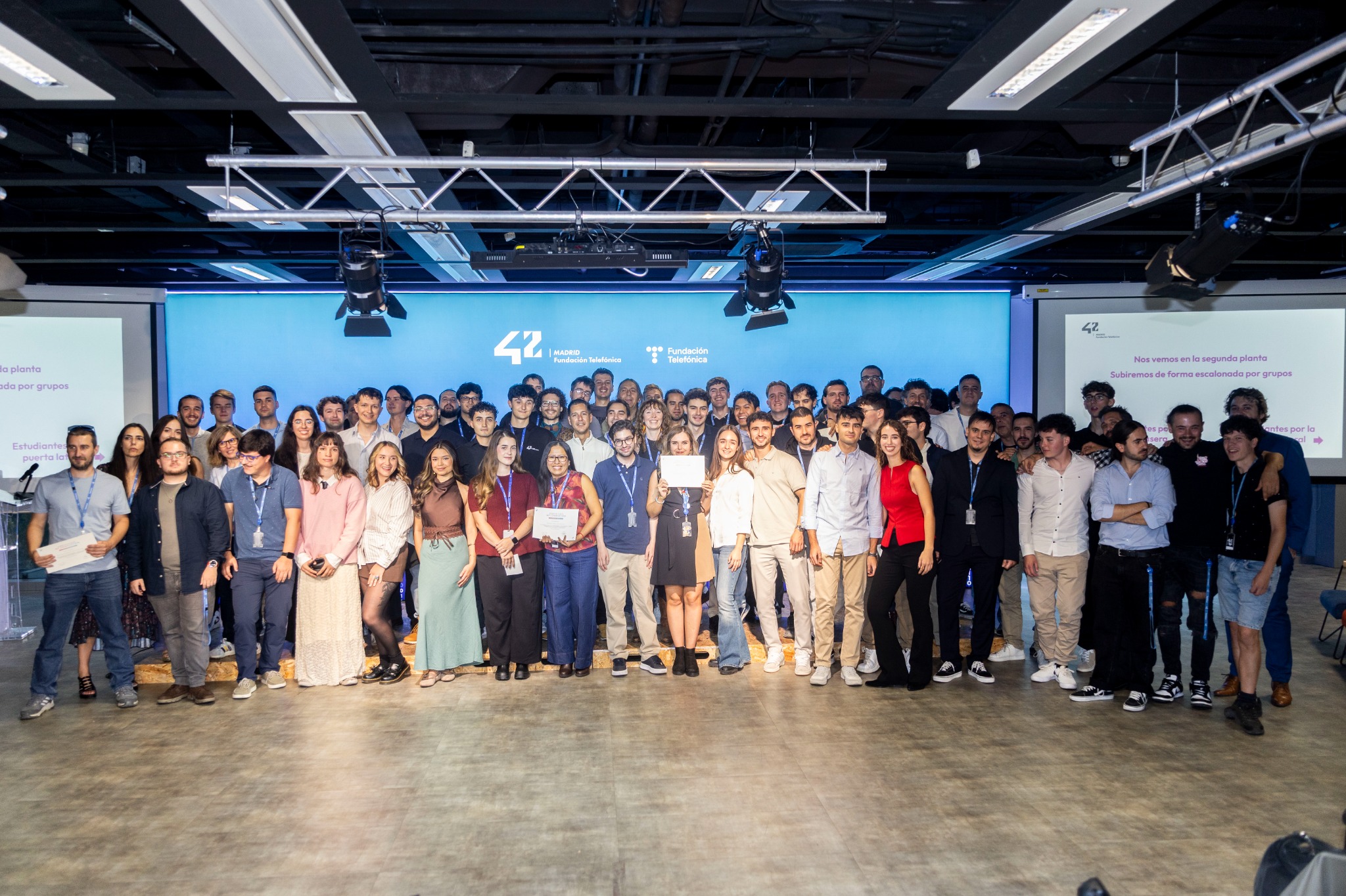 Large group posing on a stage in front of a blue backdrop with logos, some holding certificates.