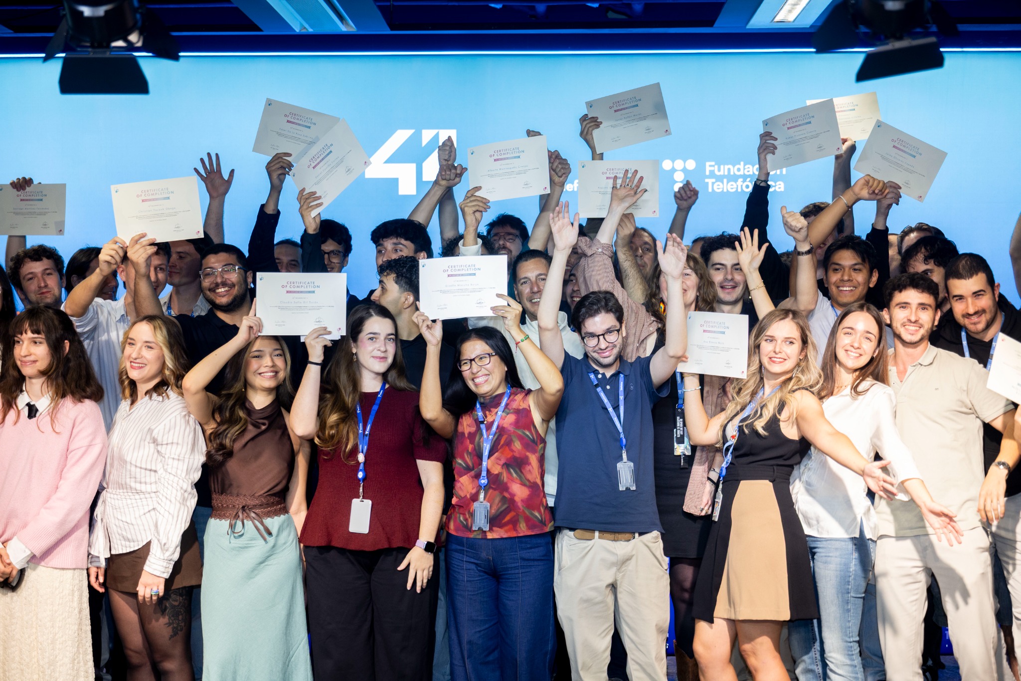 Diverse group of people on a stage, smiling and raising certificates of completion above their heads in celebration.