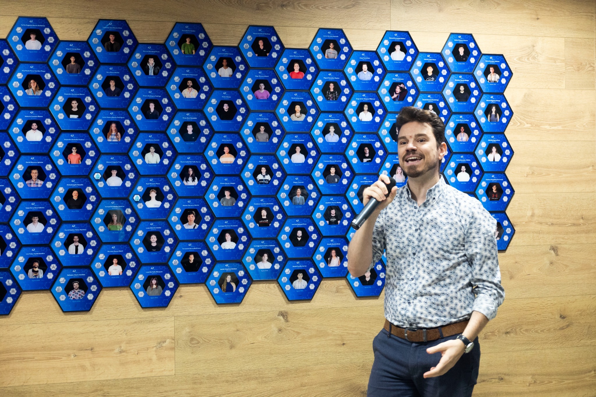 1Y8A3641_web Man with a microphone standing in front of a wall of blue hexagon plaques with photos, likely giving a talk or presentation.