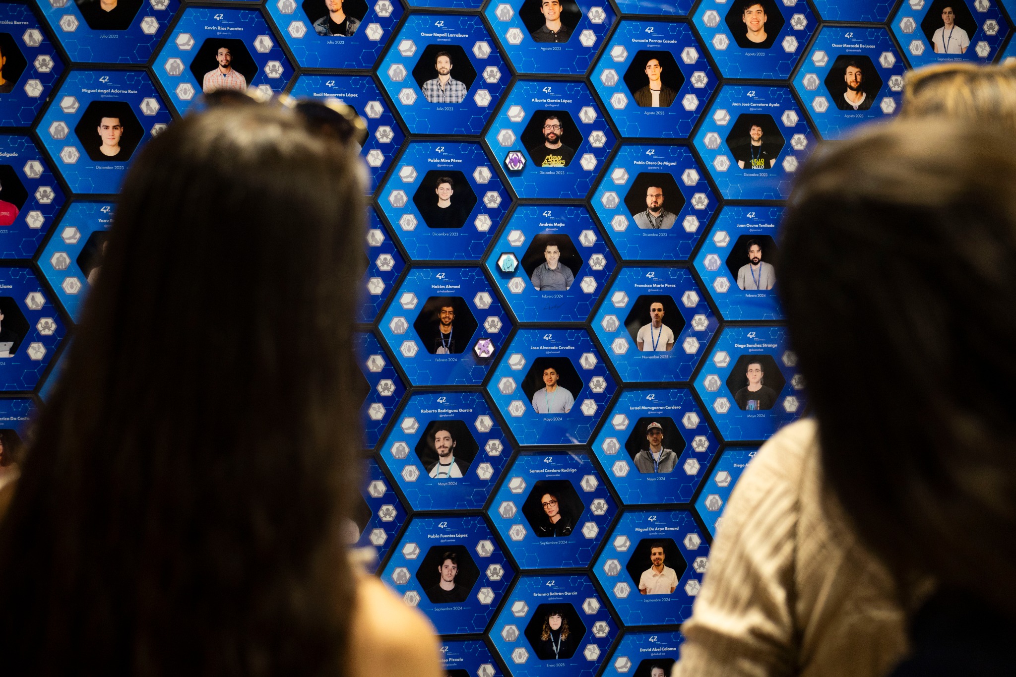 Blue wall covered with hexagonal panels showing portraits and names of people, with two blurred attendees in the foreground.