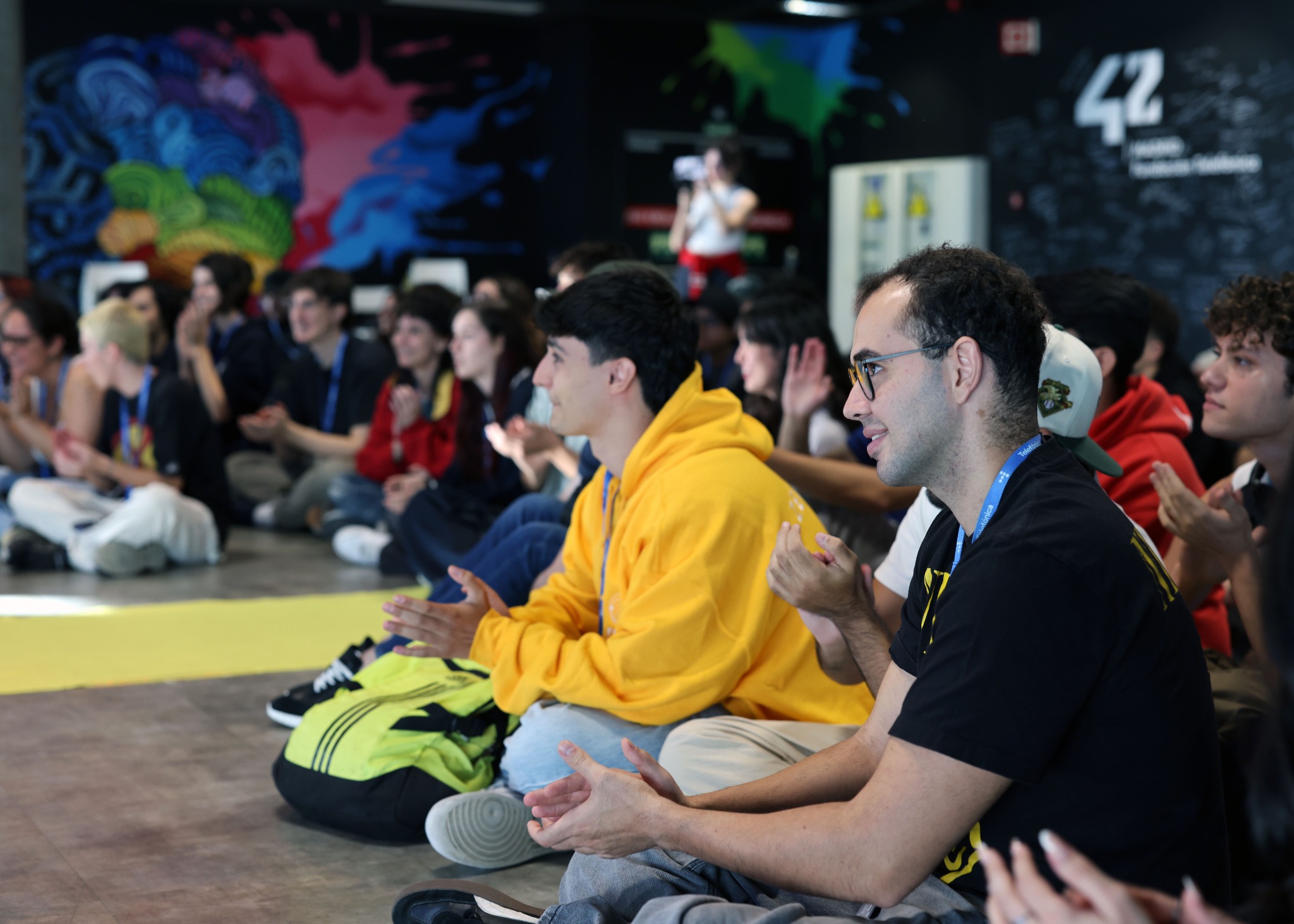 IMG_0864_web Audience of young people sitting on the floor and clapping at an indoor event, colorful mural in the background