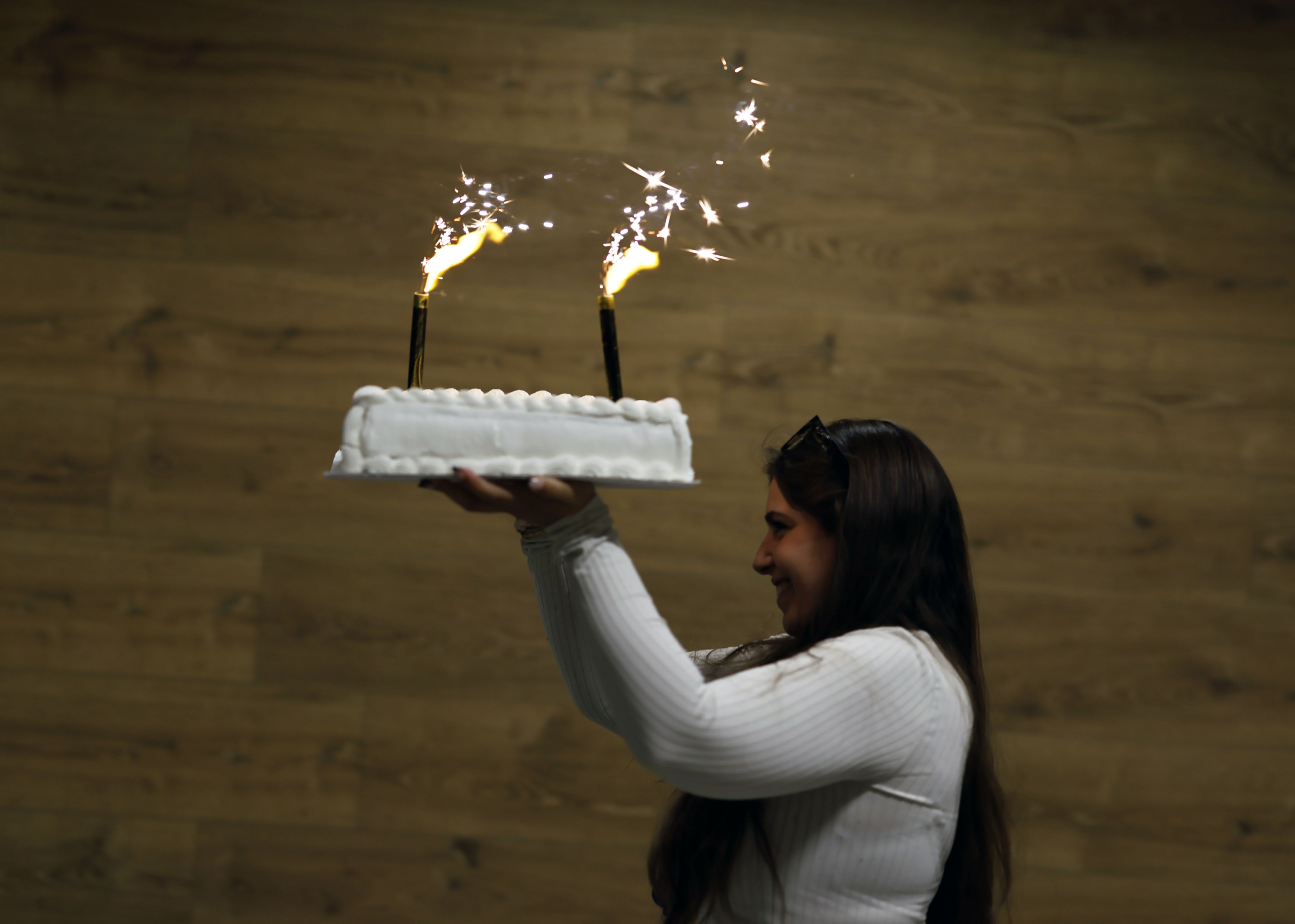 _MG_1215 Person in a white ribbed sweater holds a white frosted cake with two sparklers lit on top against a wooden wall background.